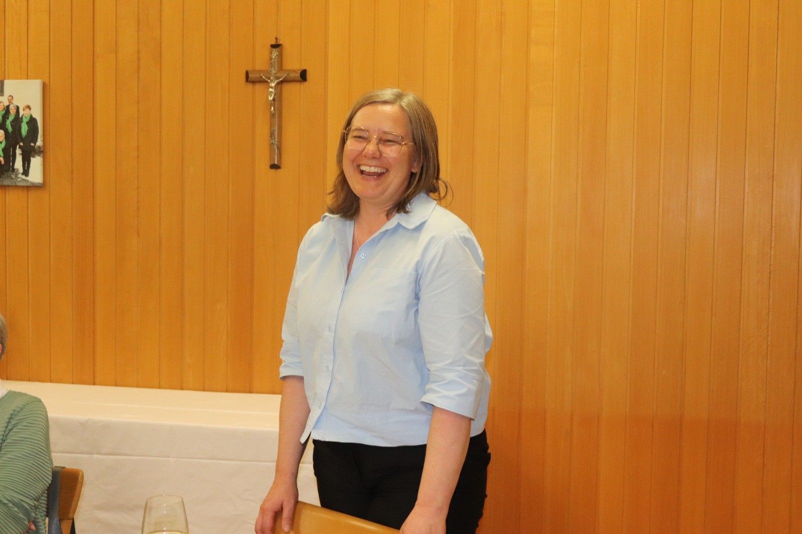 Eine lächelnde Frau mit Brille steht vor einer holzverkleideten Wand mit einem Kreuz. Sie trägt ein hellblaues Blusenhemd und schwarze Hose. Hinter ihr befindet sich ein Tisch und ein Stuhl.