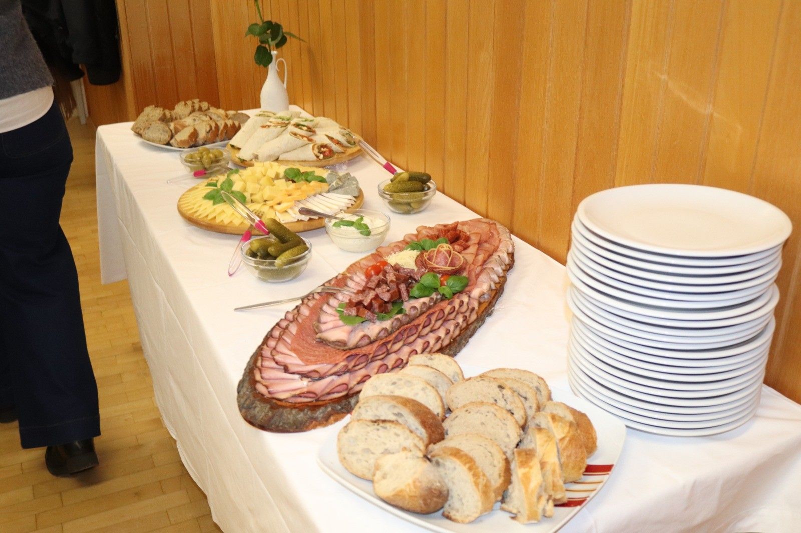 Ein Tisch mit weißer Tischdecke ist mit verschiedenen Lebensmitteln wie Fleisch, Käse, Brot und Gurken bestückt. Ein Vase mit einer Pflanze steht auf dem Tisch.