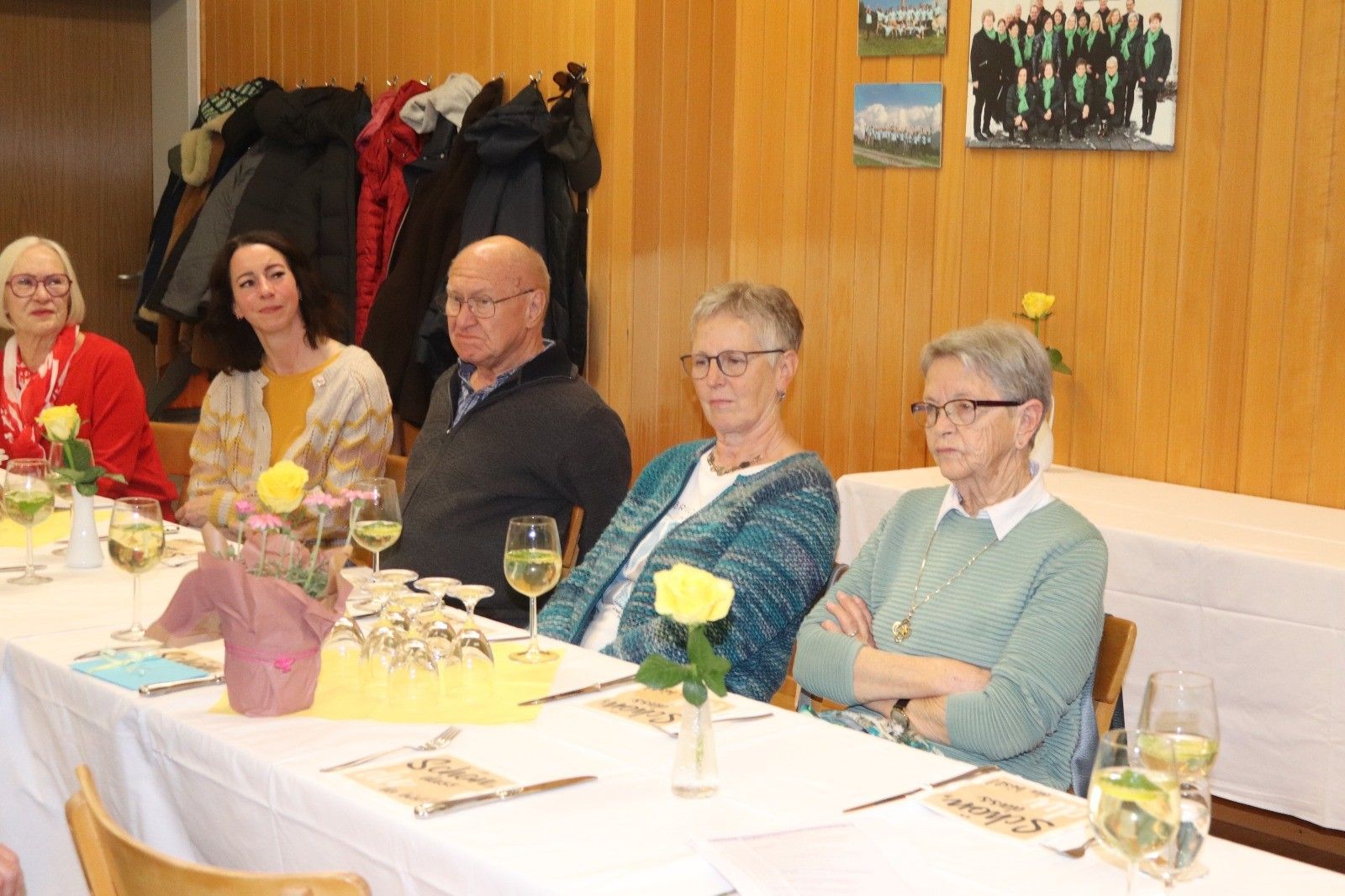 Eine Gruppe von Menschen sitzt an einem Tisch mit Weingläsern und Blumen. Eine Frau hält einen Blumenstrauß. Hinter ihnen hängen Jacken an Haken.