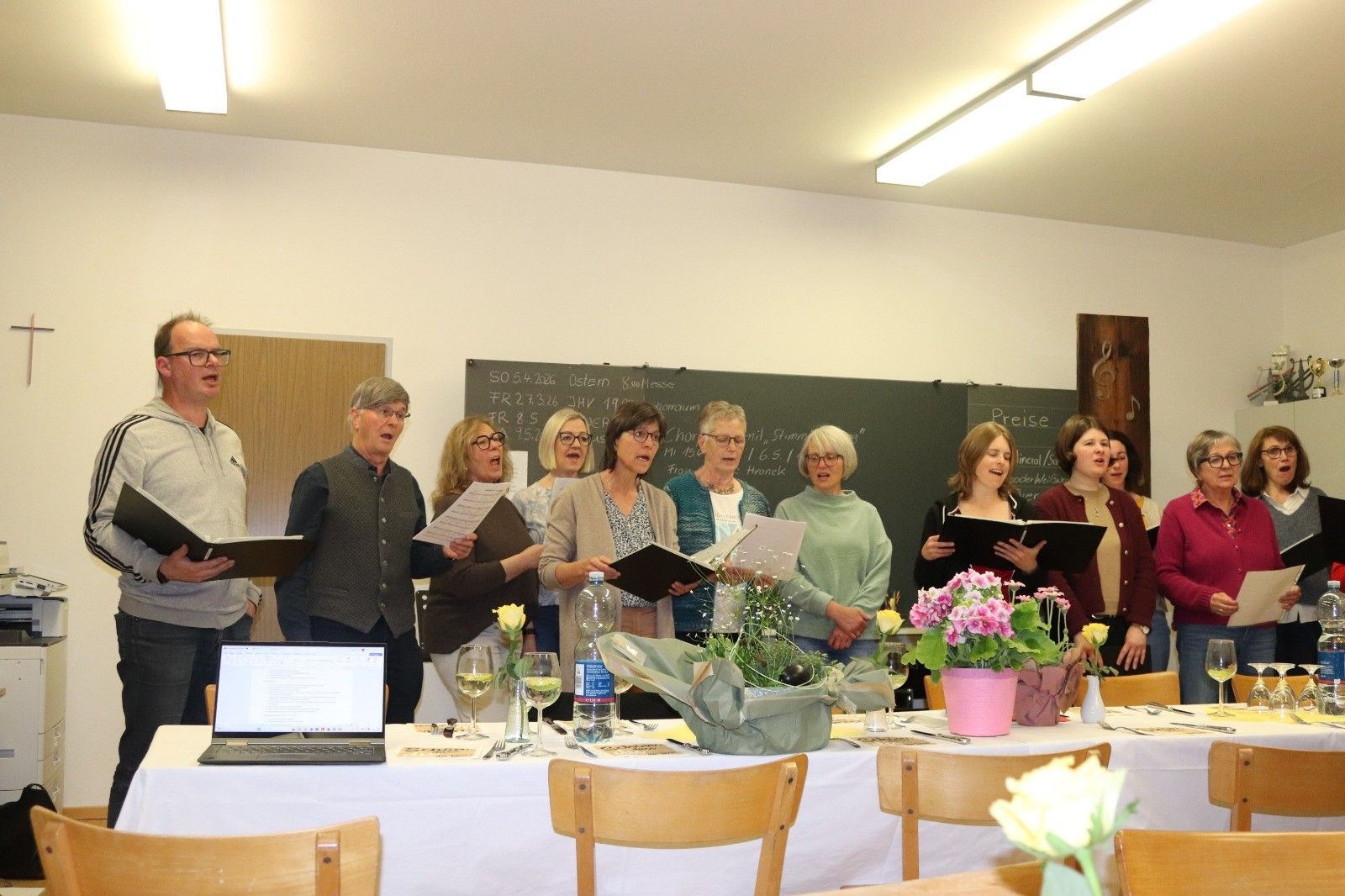 Eine Gruppe von Menschen singt mit Musikbüchern und Blumen vor einer Tafel.