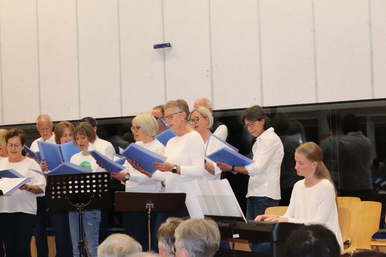 Ein Chor von Menschen in weißen Hemden singt mit blauen Broschüren in der Hand in einem Raum mit weißen Wänden und einem Klavier auf der Bühne.