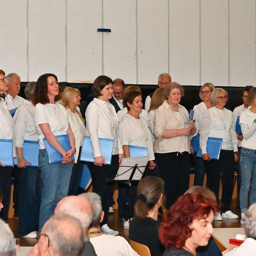 Ein Chor von Frauen steht in einer Reihe und hält blaue Ordner. Sie sind wahrscheinlich bei einer formellen Veranstaltung. Ein Publikum sitzt vor ihnen, einige tragen Brillen. Die Bühne hat eine weiße Wand mit schwarzem Hintergrund.