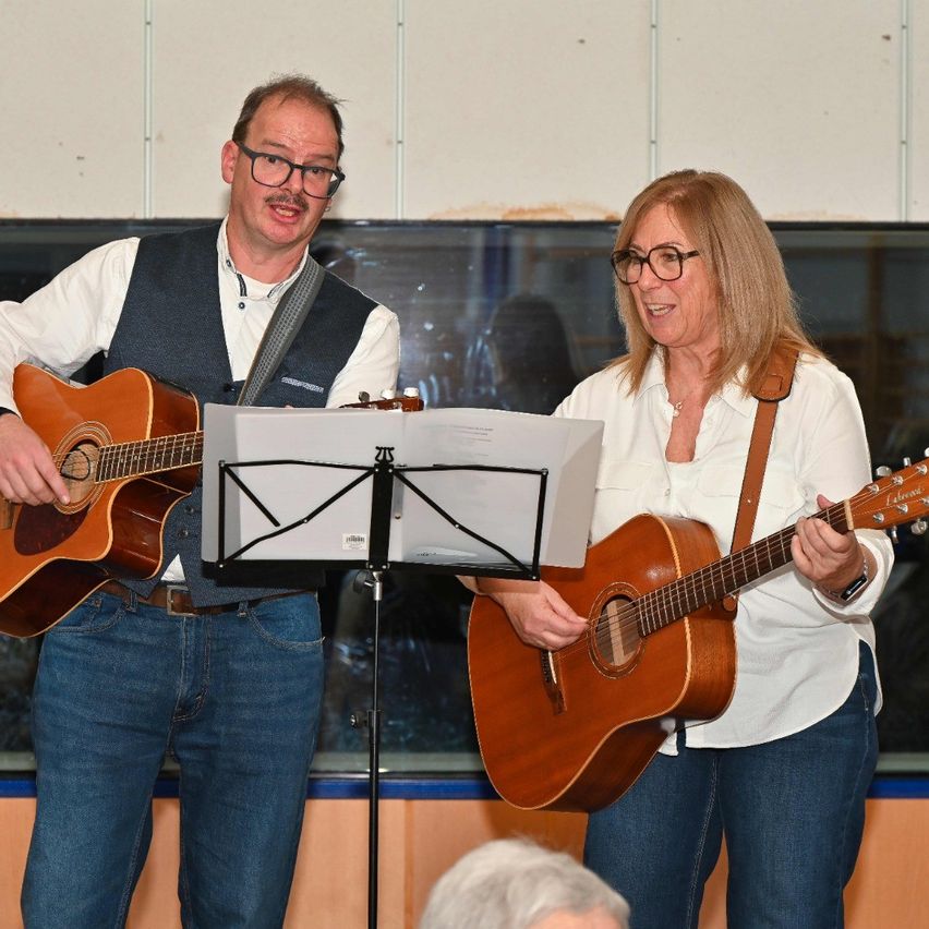 Ein Mann und eine Frau stehen nebeneinander und spielen beide Gitarre. Der Mann trägt eine Weste und eine Brille, die Frau ein weißes Hemd und eine Brille. Ein Notenständer steht zwischen ihnen.