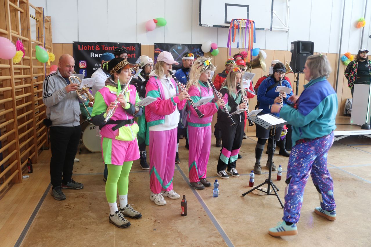 Eine Gruppe von Menschen in leuchtenden pinken und grünen Trainingsanzüge, spielen Instrumente in einer Turnhalle.