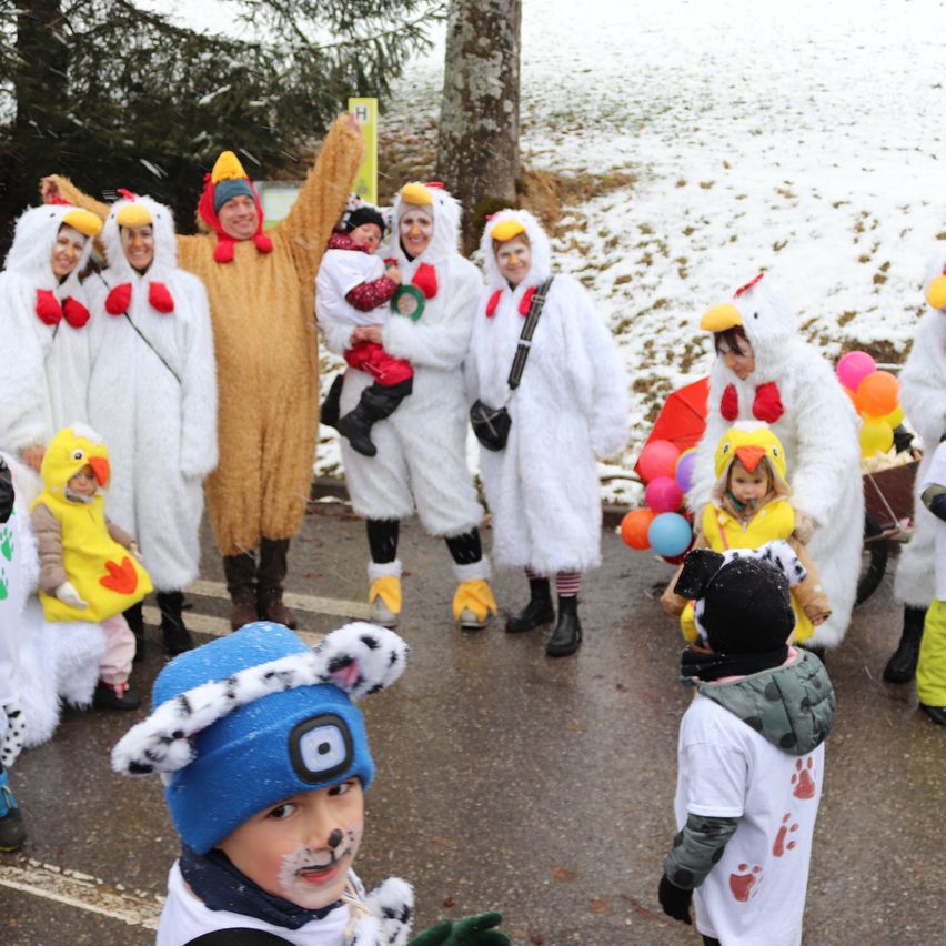 Eine Gruppe von Menschen in Hühnerkostümen, einige in Gelb und einer in Braun, stehen auf einer verschneiten Straße. Ein Kind in einem Hundekostüm steht davor.