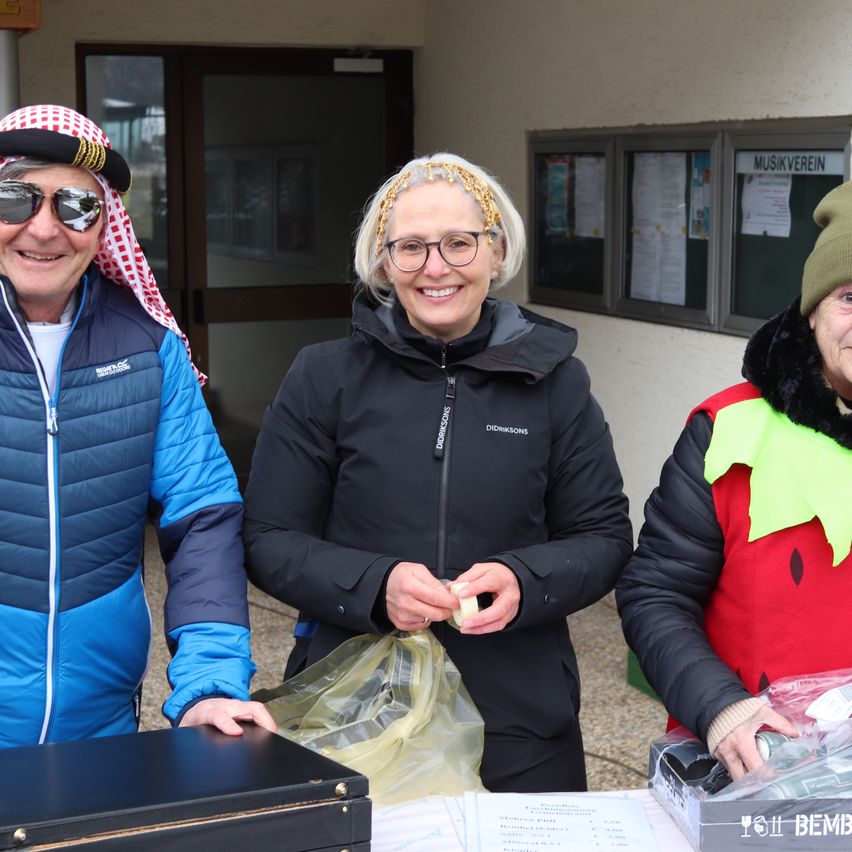 Drei Menschen stehen lächelnd draußen. Der Mann auf der linken Seite trägt ein rot-weißes Kopftuch, eine Sonnenbrille und eine blaue Jacke. Die Frau in der Mitte mit Brille hält ein kleines Objekt in der Hand. Die Frau auf der rechten Seite trägt einen grünen Hut und ein rotes und grünes Kostüm. Hinter ihnen steht ein Gebäude mit Glastüren und Fenstern.