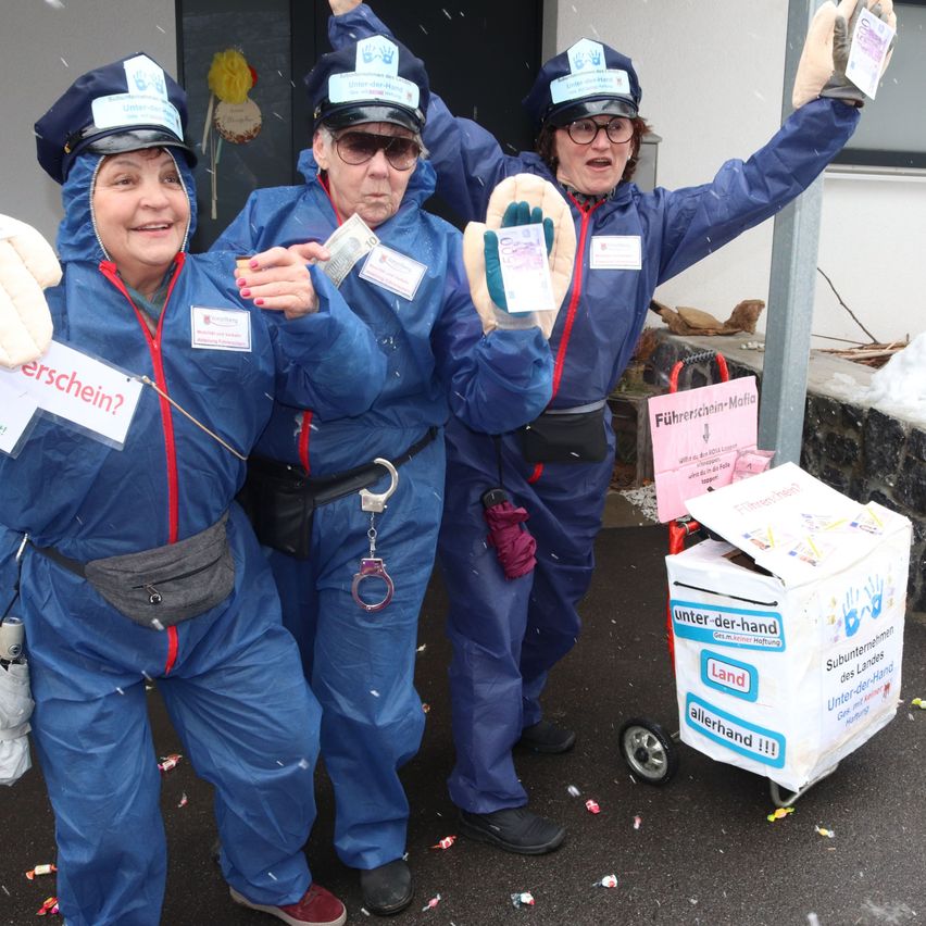 Drei Frauen in blauen Polizeiuniformen stehen zusammen, eine hält einen Geldbeutel. Sie lächeln und scheinen zu feiern. Hinter ihnen steht ein Trolley mit einem Schild, das 'Führerschein-Mafia' liest. Schnee liegt auf dem Boden.