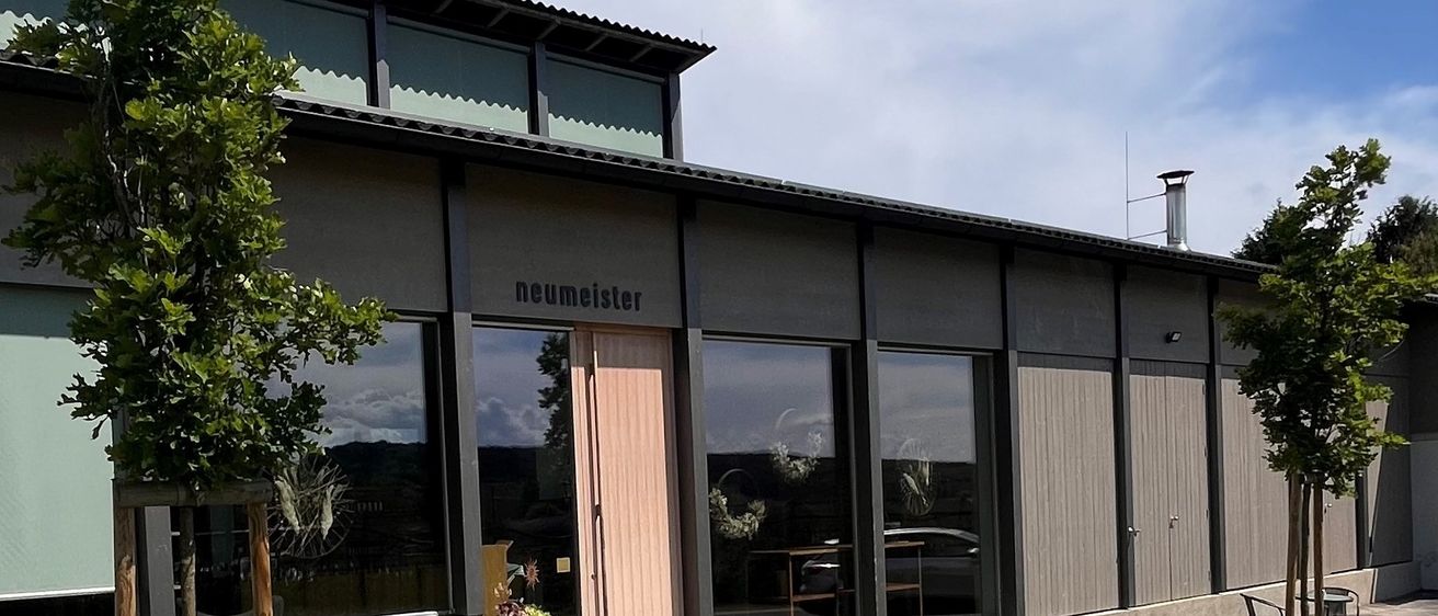 Ein modernes Gebäude mit großen Glasfenstern und einer rosa Tür. Es hat einen Gehweg mit einem kleinen Garten und Topfpflanzen davor. Das Gebäude hat einen Schornstein und ein Schild mit der Aufschrift 'neumeister'. Der Himmel ist bewölkt.