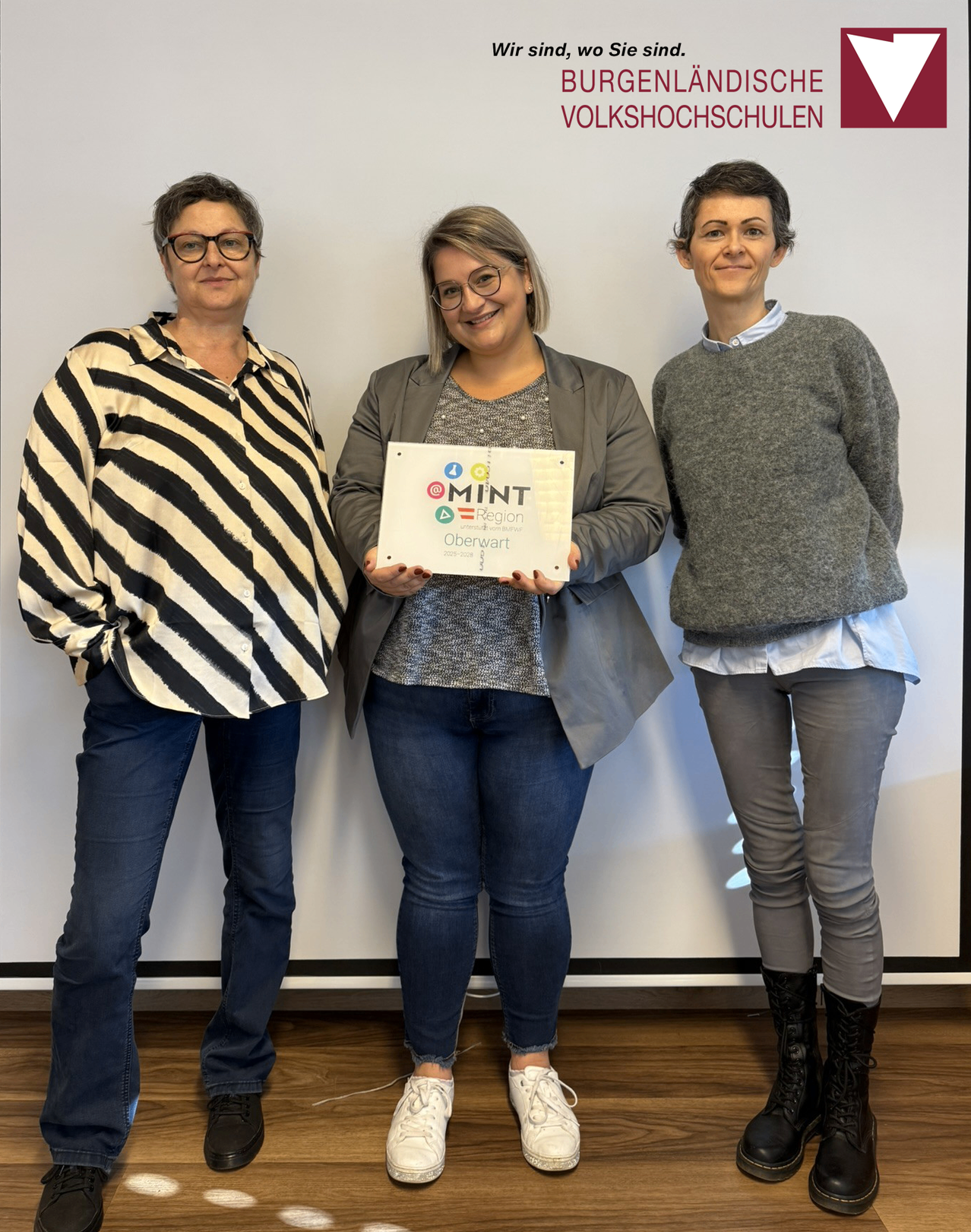 Drei Frauen stehen lächelnd nebeneinander und halten ein Zertifikat in der Hand, vor einem weißen Hintergrund. Die Frau in der Mitte hält ein Zertifikat mit 'MINT' darauf.
