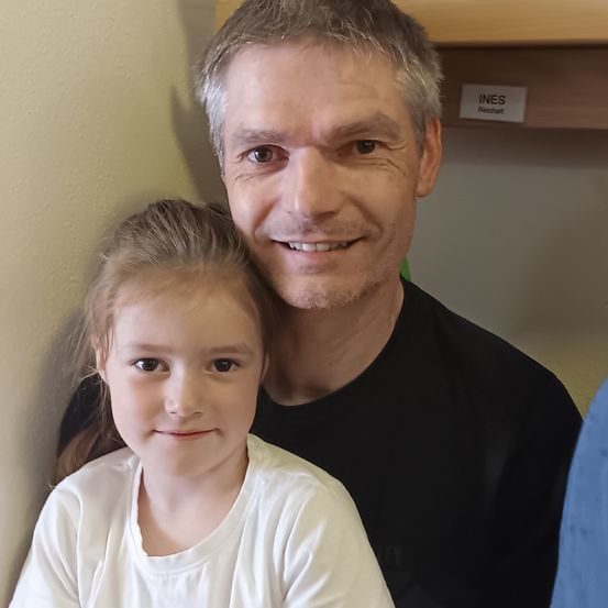 Ein lächelnder Mann in einem schwarzen Shirt und ein junges Mädchen in einem weißen Shirt posieren für ein Foto vor einer gelben Wand.