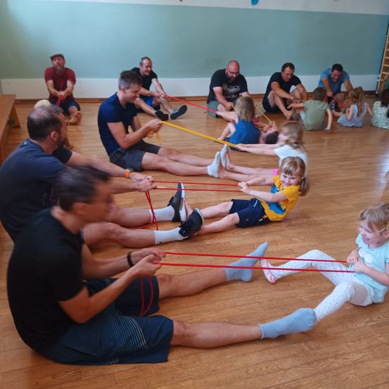 Eine Gruppe von Erwachsenen und Kindern trainiert mit Widerstandsbändern auf einem Holzboden, einige sitzen auf dem Boden und einige stehen.