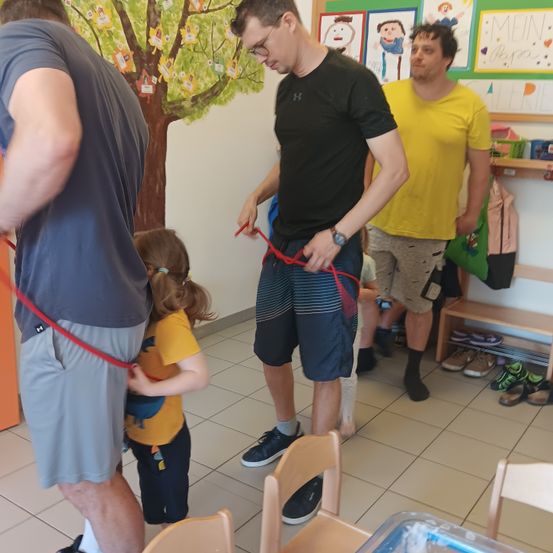 Drei Männer und ein junges Mädchen befinden sich in einem Raum. Ein Mann befestigt einen Gurt am Mädchen. An der Wand befindet sich eine Baummalerei und Schuhe stehen auf einem Regal.