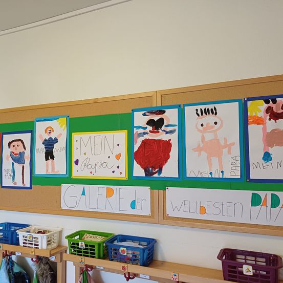 Eine Klassenzimmerwand zeigt verschiedene bunte Kinderzeichnungen, jeweils mit 'Mein Papa' beschriftet. Die Wand ist mit grünen Rändern dekoriert. Unten stehen Regale mit verschiedenen Aufbewahrungskörben.