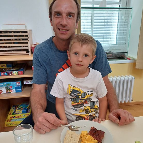 Ein Mann und ein junger Junge sitzen lächelnd am Tisch. Der Junge hält einen Teller mit Eiern, Brot und Speck. Dahinter steht ein Regal mit Spielzeug und ein Fenster mit Jalousien.