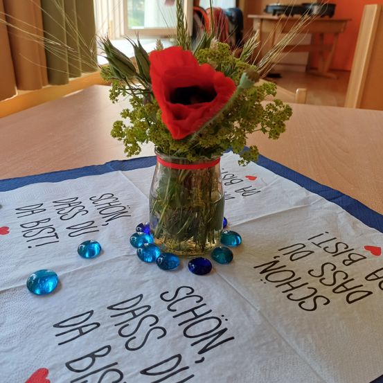 Ein Blumenarrangement mit einer roten Mohnblume und grünen Blättern in einem Glasvase auf einem Tisch. Blaue Perlen und ein Tuch mit Worten sind auf dem Tisch.