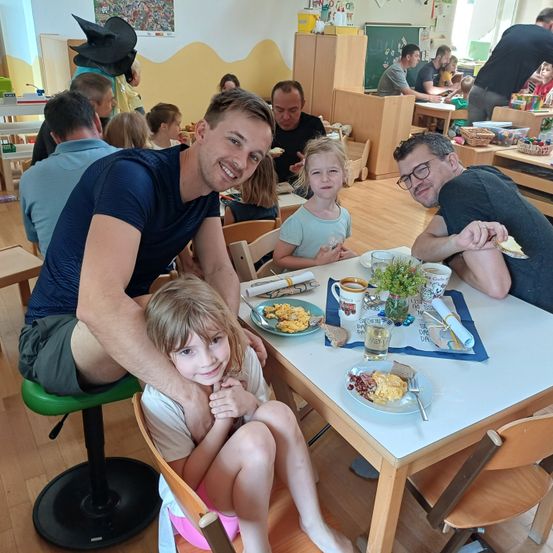 Eine vierköpfige Familie sitzt um einen Tisch in einem Klassenzimmer und lächelt. Zwei Erwachsene und zwei Kinder, von denen eines auf einem Hocker sitzt. Der Tisch hat Essen, Getränke und Besteck. Im Hintergrund sitzen und stehen weitere Personen.