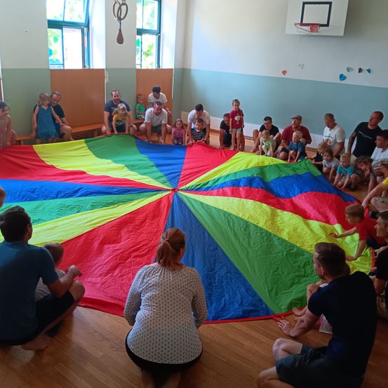 Eine Gruppe von Menschen, einschließlich Kindern, versammelt sich um einen großen, bunten Fallschirm auf einem Holzboden. Einige Erwachsene sitzen, während andere stehen oder knien. Der Raum hat weiße Wände und einen Basketballkorb im Hintergrund.