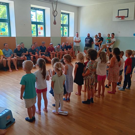 Eine Gruppe von Erwachsenen und Kindern versammelt sich in einem Raum. Einige Erwachsene sitzen auf einer Bank, während Kinder in einem Kreis stehen. Eine Basketballkorb befindet sich an der Wand.