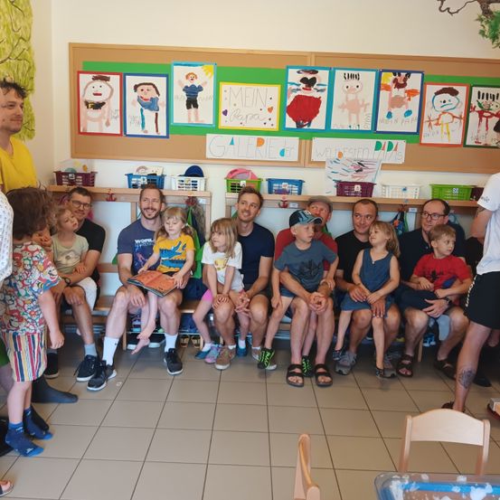 Eine Gruppe von Menschen, einschließlich Erwachsener und Kinder, sitzt auf einer Bank in einem Klassenzimmer. Ein Mann liest den Kindern ein Buch vor. Dahinter befinden sich Regale mit Körben und Zeichnungen an der Wand.