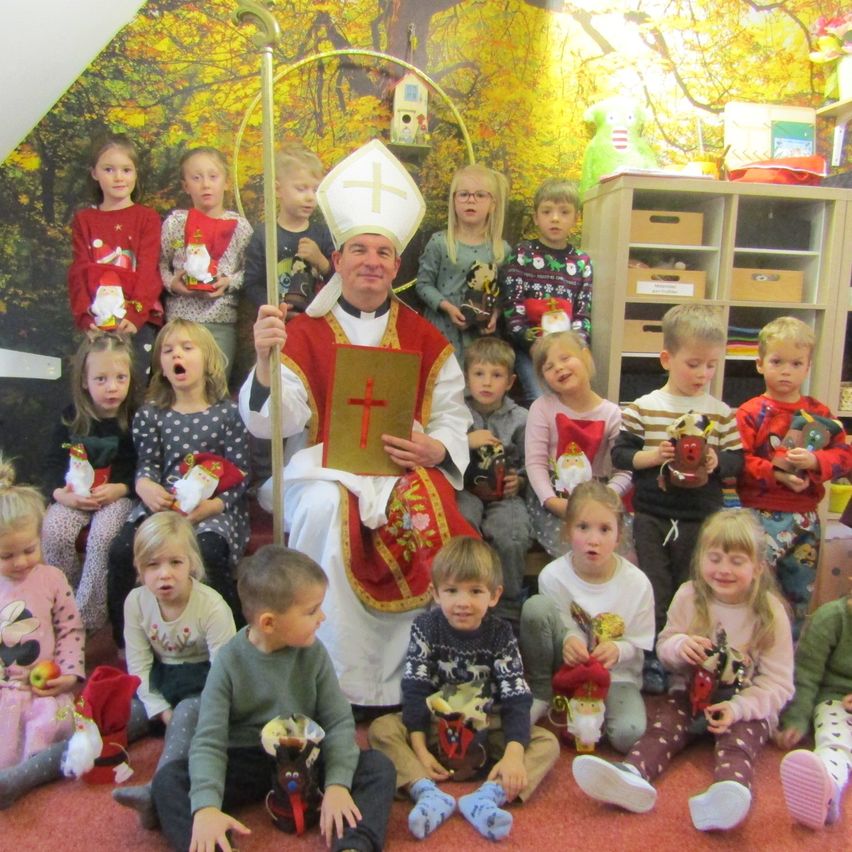 Eine Gruppe von Kindern mit ihrem Lehrer in festlicher Kleidung, posiert für ein Foto mit einem Priester vor einer Kulisse, die Herbstbäume darstellt.