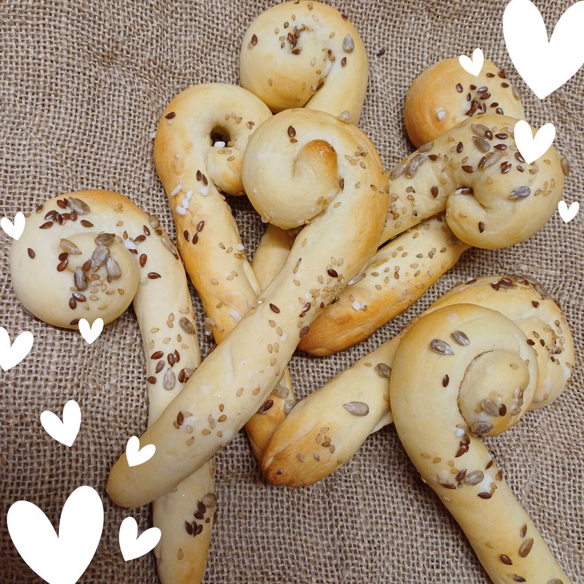 Eine Sammlung von Brezeln mit Samen auf einem Jutehintergrund. Die Brezeln sind goldbraun und in verschiedene Formen verdreht. Herzen sind um die Brezeln verstreut.