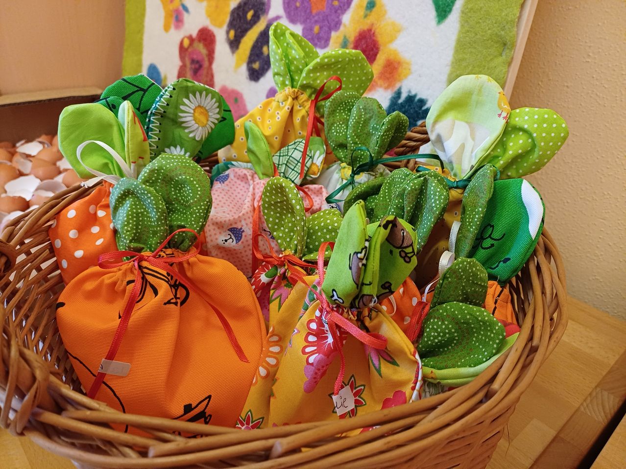 Ein Korb voller bunter, handgenähter Stoffbeutel mit Bändern, die jeweils wie ein Hase oder eine Karotte gestaltet sind. Sie sind in einem Kreismuster angeordnet, mit einem Blumenhintergrund hinter dem Korb.