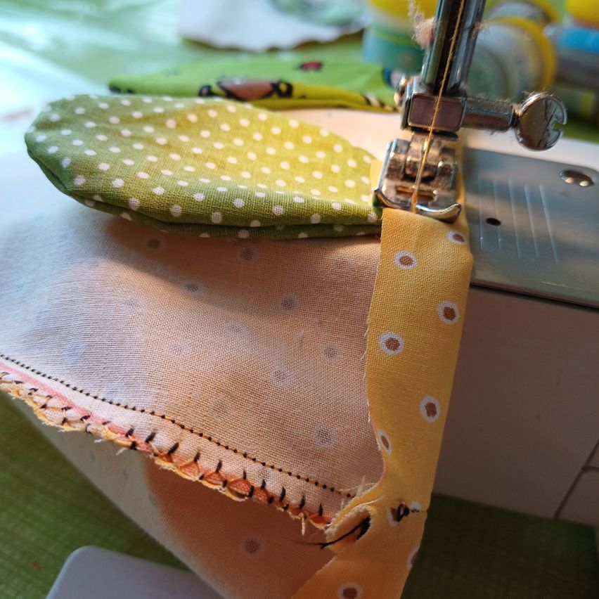 Eine Nähmaschine näht ein Stück Stoff mit Punktemuster auf einer grünen Oberfläche. Ein gelber Stoff mit Punktemuster ist ebenfalls sichtbar.