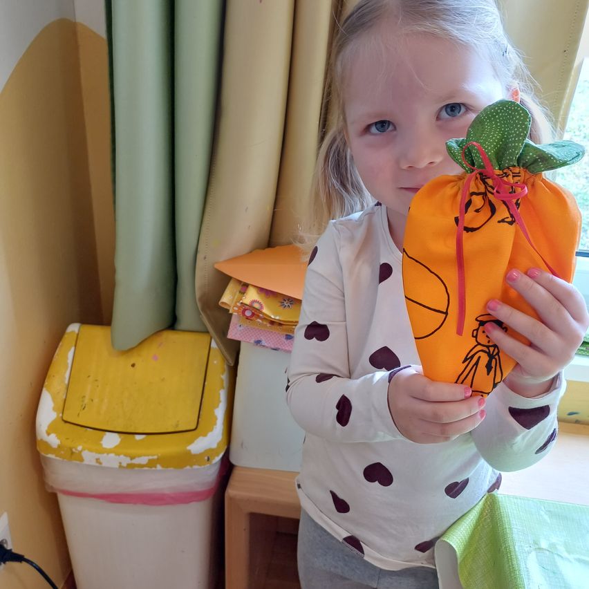 Ein junges Mädchen mit blonden Haaren hält eine orange Tasche mit grünem Oberteil und rotem Band. Sie trägt ein weißes Langarm-Shirt mit Punkten. Hinter ihr steht ein Mülleimer und ein Vorhang.