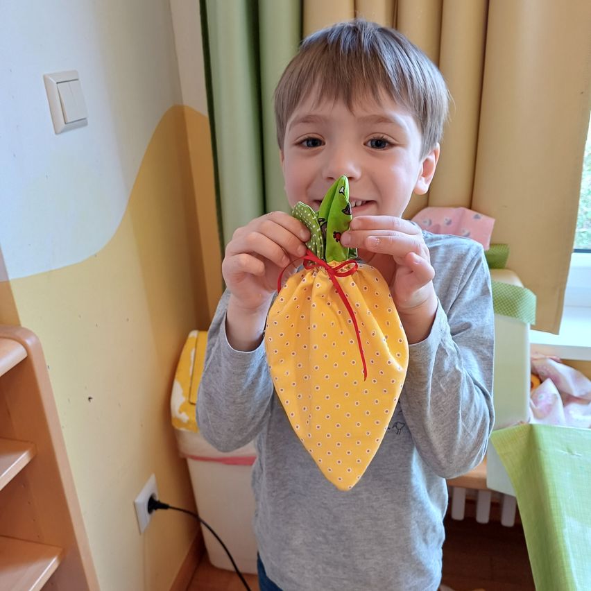 Ein kleiner Junge hält einen gelben Beutel mit einem roten Faden und lächelt für ein Foto. Dahinter ist ein grüner Vorhang, ein weißer Mülleimer und eine gelb gemusterte Wand.