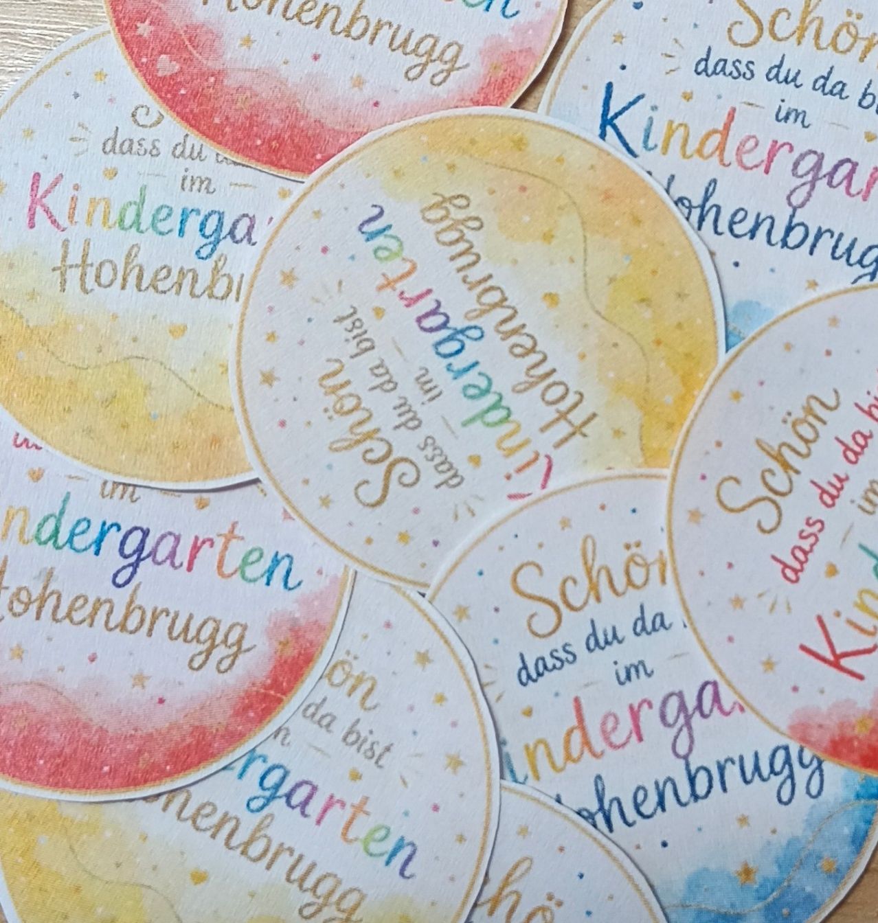 A collection of colorful circular stickers with handwritten text in various languages, including 'Kindergarten Hohenbrugg' and 'Schön dass du da bist'. The stickers have a watercolor background and are arranged in a cluster.