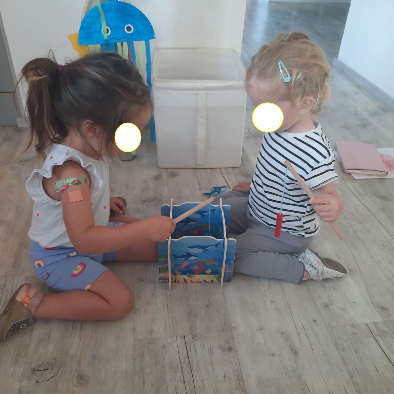 Zwei kleine Mädchen sitzen auf einem Holzboden, eine spielt ein Spielzeugtrommel, während die andere einen Drumstick hält. Ein weißer Aufbewahrungskasten steht hinter ihnen, und ein blauer Quallen-Papierbastelwerk hängt an der Wand.