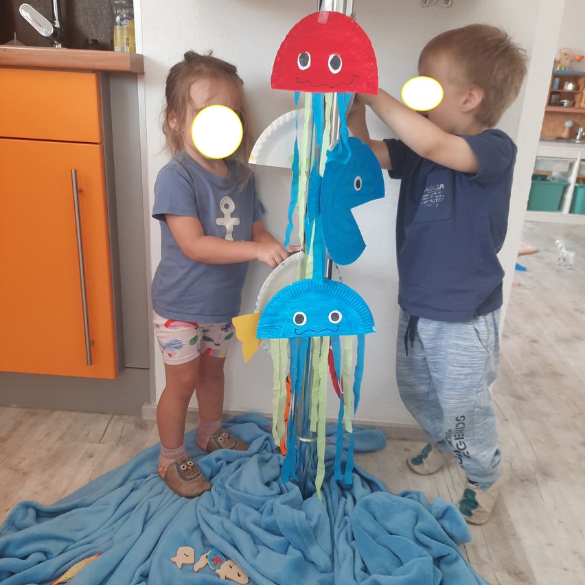 Zwei Kinder stehen in einem Raum, eines hält eine Quallen-Bastelei aus Papiertellern und Bändern, während das andere sie justiert. Ein blauer Teppich liegt auf dem Boden.
