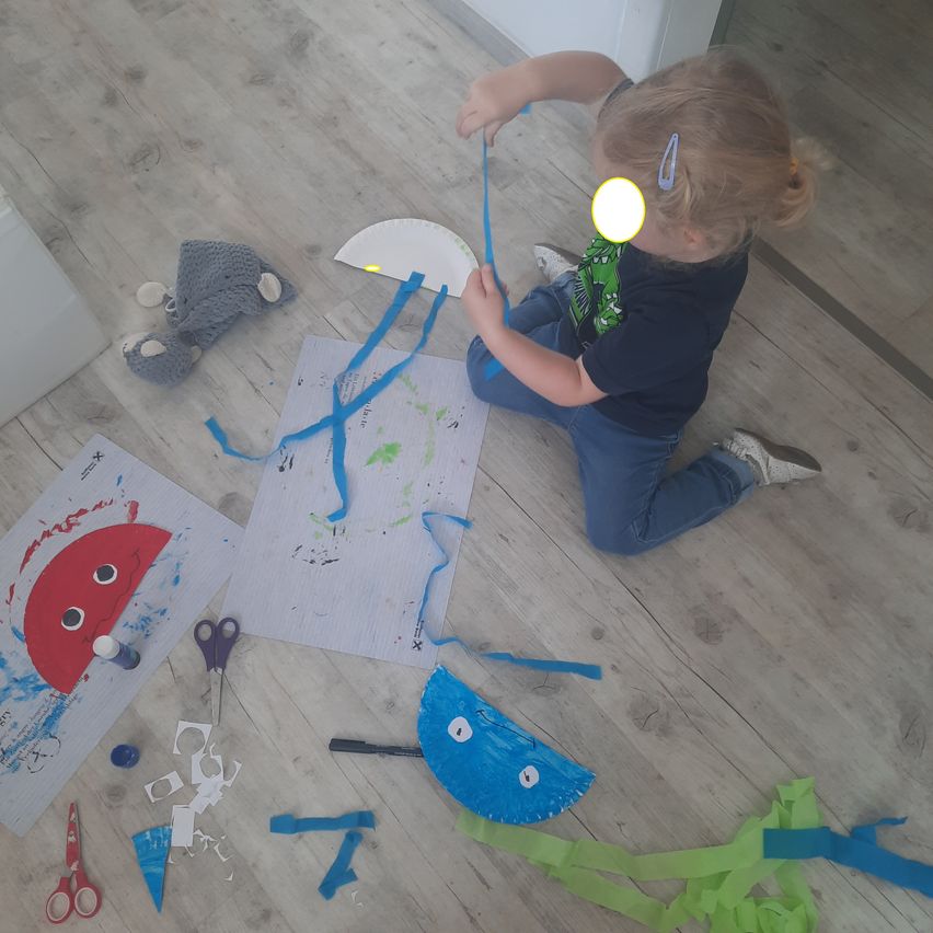 Ein kleines Mädchen sitzt auf dem Boden, umgeben von Bastelmaterialien. Sie hält eine blaue Schleife und neben ihr liegt ein blauer Papierfisch. Andere Materialien umfassen Scheren, Papierausschnitte und ein graues Kuscheltier. Der Boden ist aus Holz und der Bereich ist gut beleuchtet.