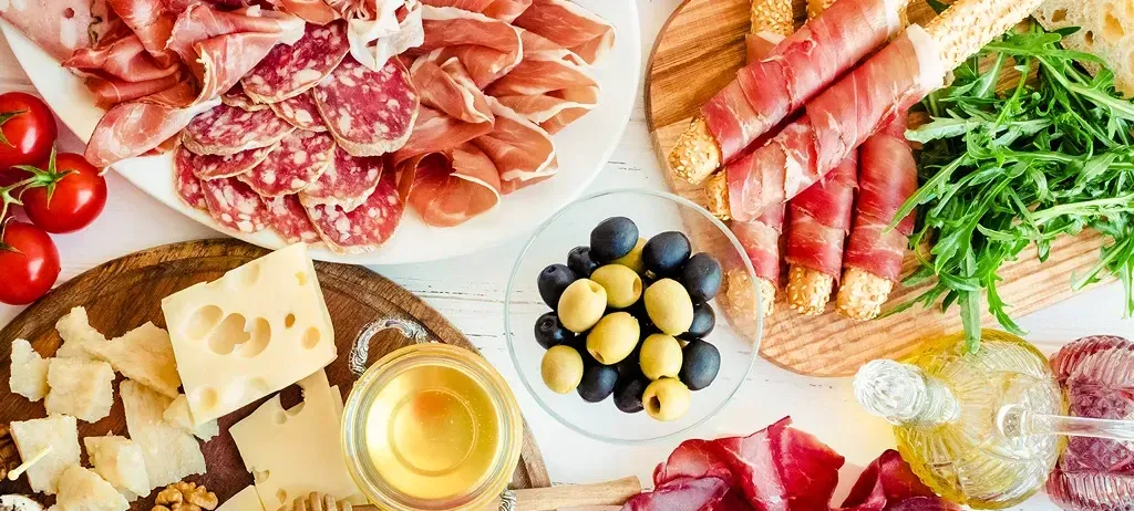 Eine Auswahl an Fleisch, Käse, Oliven, Tomaten und Kräutern auf weißem Hintergrund. Ein Honigglas, eine Olivenölflasche und eine Zitronenscheibe begleiten das Angebot.