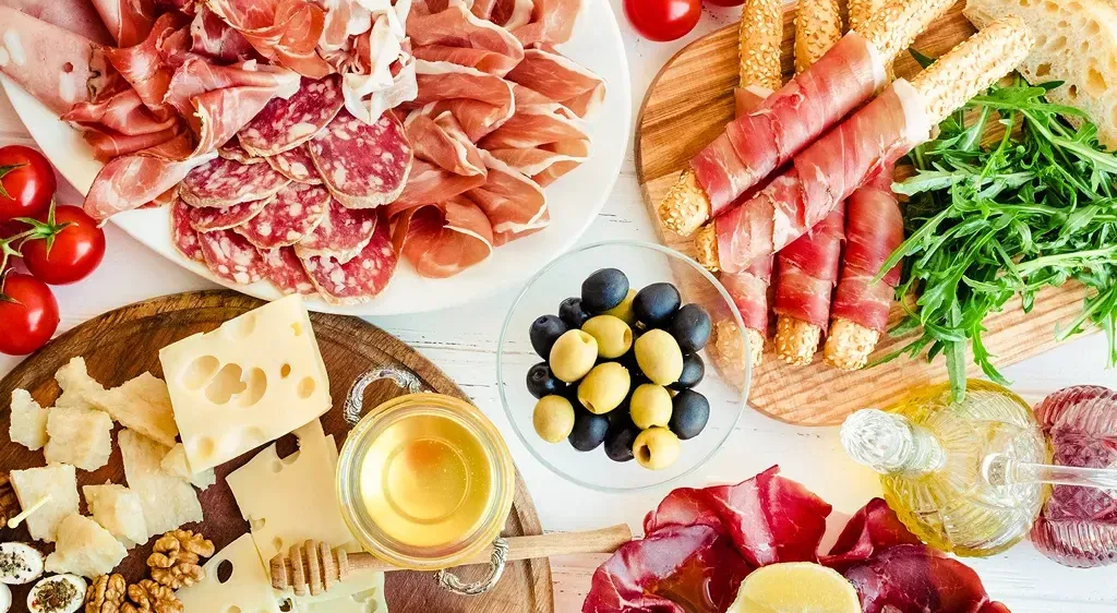 Eine Auswahl an Fleisch, Käse, Oliven, Tomaten und Kräutern auf weißem Hintergrund. Ein Honigglas, eine Olivenölflasche und eine Zitronenscheibe begleiten das Angebot.