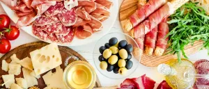Eine Auswahl an Fleisch, Käse, Oliven, Tomaten und Kräutern auf weißem Hintergrund. Ein Honigglas, eine Olivenölflasche und eine Zitronenscheibe begleiten das Angebot.