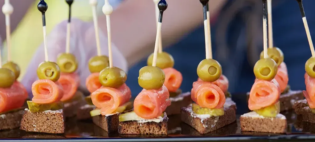 Ein Tablett hält mehrere mundgerechte Vorspeisen, jeweils mit einem Stück Lachs, einer grünen Olive und einer Scheibe Brot, alles von Zahnstochern gehalten.
