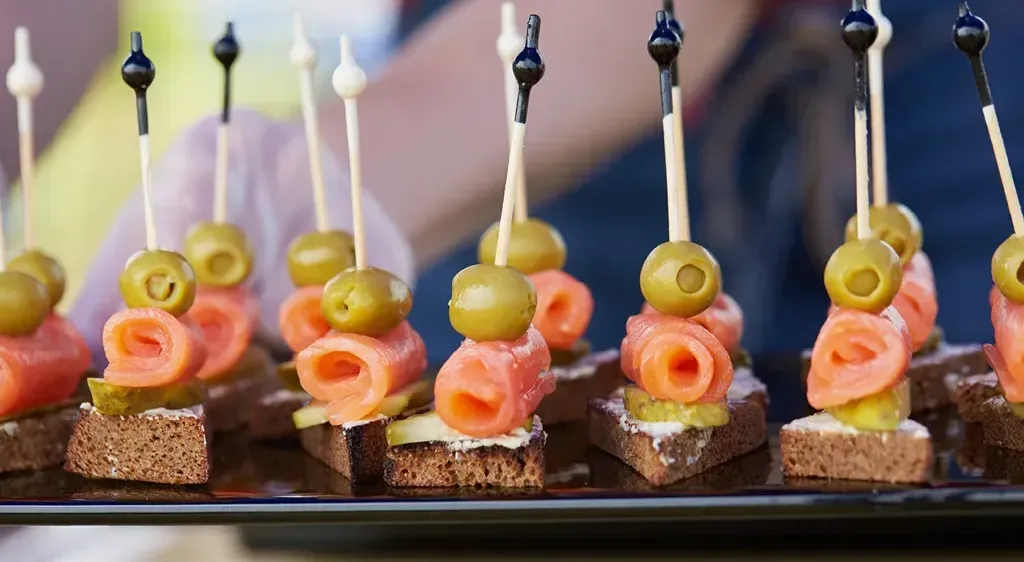 Ein Tablett hält mehrere mundgerechte Vorspeisen, jeweils mit einem Stück Lachs, einer grünen Olive und einer Scheibe Brot, alles von Zahnstochern gehalten.