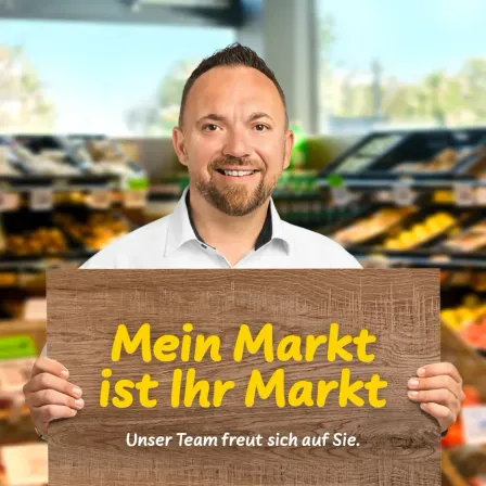 Ein Mann in einem weißen Hemd hält ein hölzerne Schild mit gelbem Text, das 'Mein Markt ist Ihr Markt' sagt und lächelt in die Kamera in einem Supermarkt.