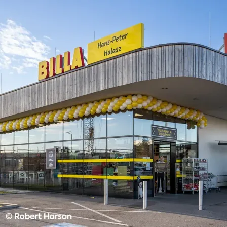 Die Vorderseite eines BILLA-Geschäfts mit gelber und weißer Luftballondekoration. Ein Mann steht am Eingang, und ein Hund ist neben ihm. Das Geschäft hat eine Glaswand und Tür, und ein Schild steht Willkommen Billa Zoodorf.