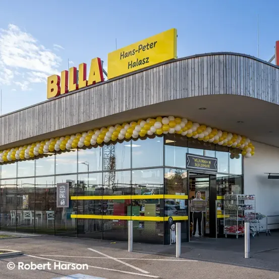 Die Vorderseite eines BILLA-Geschäfts mit gelber und weißer Luftballondekoration. Ein Mann steht am Eingang, und ein Hund ist neben ihm. Das Geschäft hat eine Glaswand und Tür, und ein Schild steht Willkommen Billa Zoodorf.