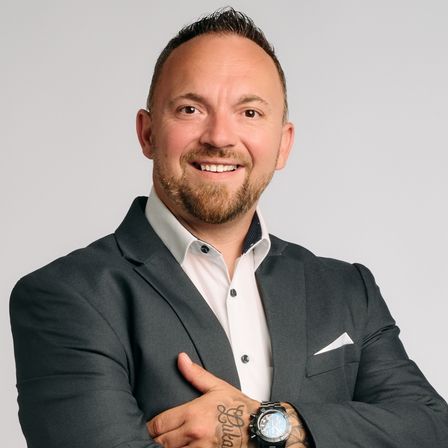 Ein Mann mit Bart und Tattoos posiert selbstbewusst mit verschränkten Armen, trägt einen dunklen Anzug, weißes Hemd und eine Uhr.