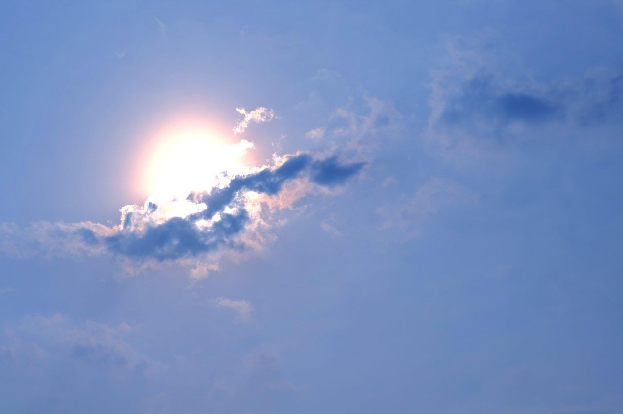 Eine strahlende Sonne scheint durch dünnflächige Wolken in einem klaren blauen Himmel. Die Sonnenstrahlen werfen ein warmes Leuchten auf die Wolken.