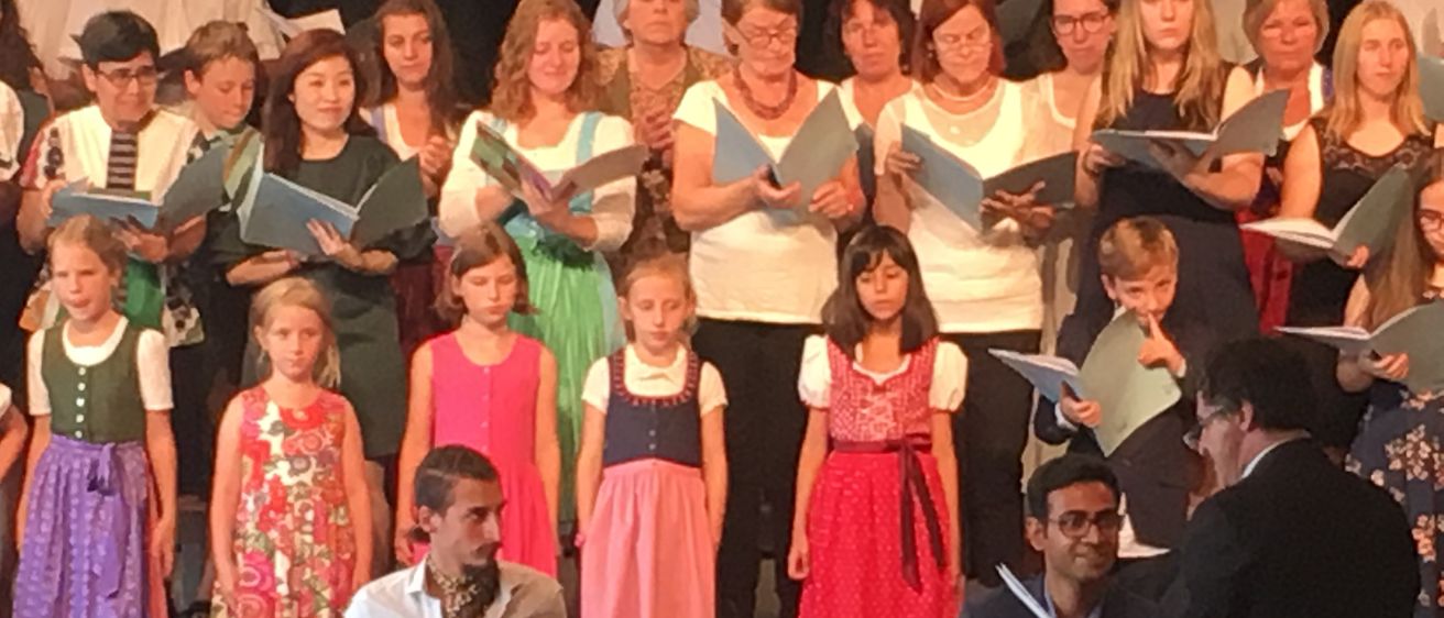 Ein Chor aus Erwachsenen und Kindern mit blauen Liederbüchern steht auf der Bühne, während zwei Männer vor ihnen Trommeln spielen. Sie veranstalten ein musikalisches Ereignis.