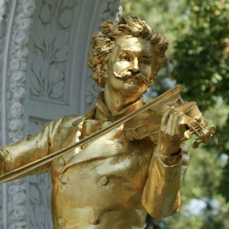 Eine goldene Statue eines Mannes mit Schnurrbart, der eine Geige spielt, steht vor einem Gebäude mit kunstvollen Verzierungen.