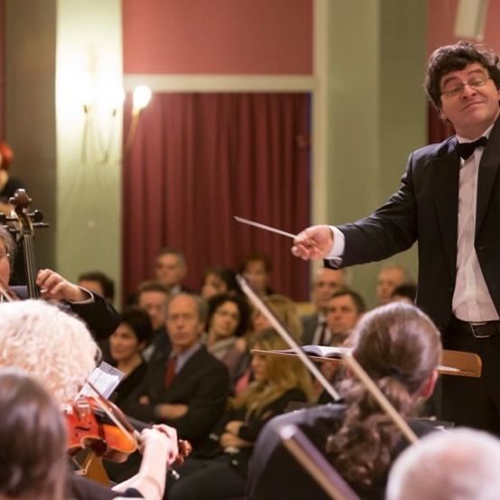 Ein Dirigent leitet ein Orchester in einem Konzertsaal, mit Musikern, die Violinen und Violoncelli spielen, und einem Publikum im Hintergrund.