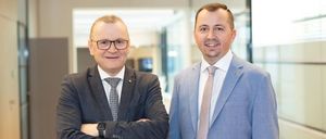 Zwei Männer in Anzügen und Krawatten stehen in einem Büro, lächeln in die Kamera, einer mit verschränkten Armen, der andere mit den Händen in den Taschen.