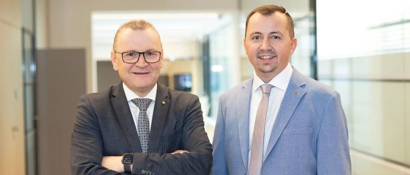 Zwei Männer in Anzügen und Krawatten stehen in einem Büro, lächeln in die Kamera, einer mit verschränkten Armen, der andere mit den Händen in den Taschen.