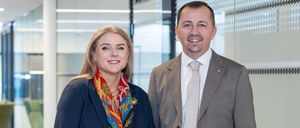 Ein Mann und eine Frau in Geschäftskleidung stehen gemeinsam in einem modernen Büroflur. Die Frau trägt einen blauen Blazer und einen bunten Schal, während der Mann einen grauen Anzug und eine Krawatte trägt. Sie lächeln beide und schauen direkt in die Kamera.