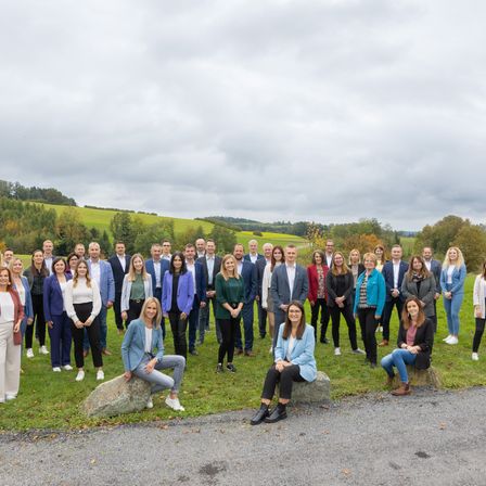 Eine große Gruppe von Geschäftsleuten, gekleidet in Geschäftskleidung, posiert für ein Foto auf einer Wiese, mit einem Schotterweg und grünen Hügeln im Hintergrund.